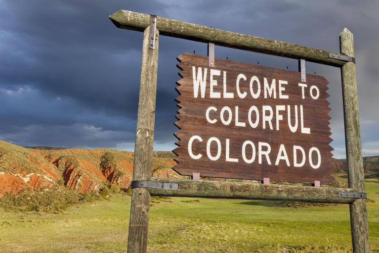 Large sign that reads "Welcome To Colorful Colorado" hangs over green grass and a vibrant, cloudy sky