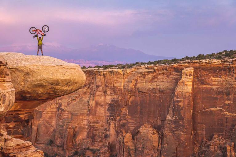 A mountain biker holds his bike over his head on the top of a large rock over looking a canyon in the Colorado desert. Photo by Dan Holz.
