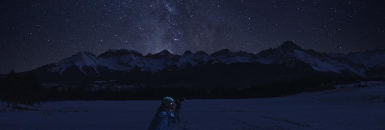 Man looking up at the stars using a telescope with mountains in the background.
