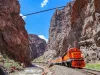 An orange train passes through a deep canyon on a sunny day.